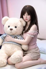 full body sex doll seated on bed holding oversized teddy bear in mid shot composition with soft pastel indoor lighting and character focus